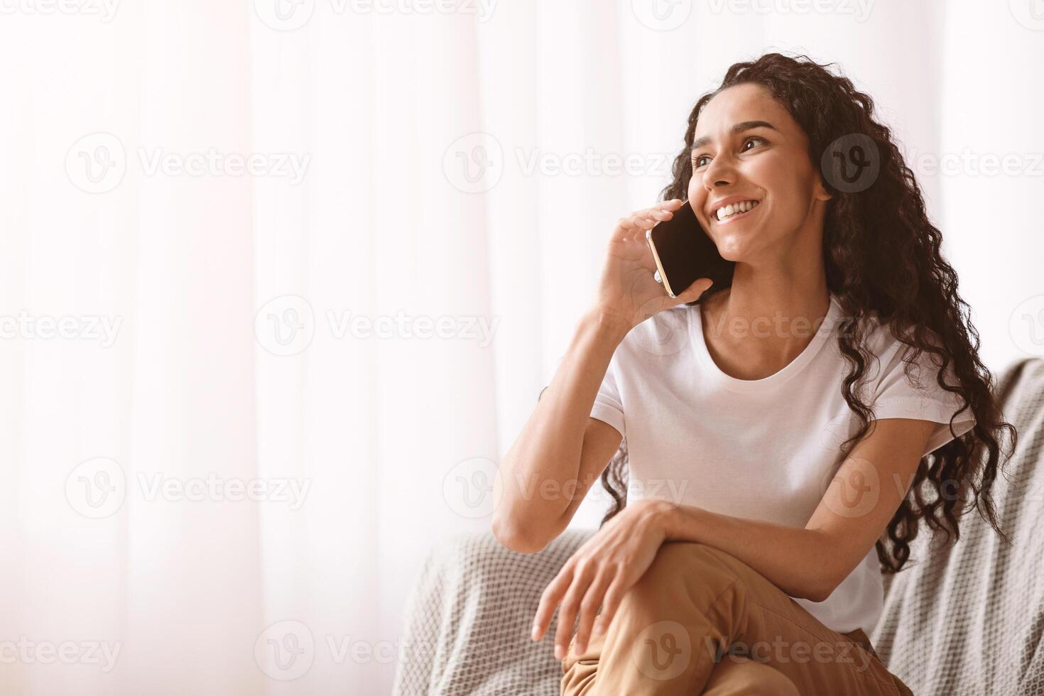 Happy young brunette lady with long curly hair in casual sitting by window at home, cheerfully talking on mobile phone with friends or lover, copy space. Communication during isolation concept photo