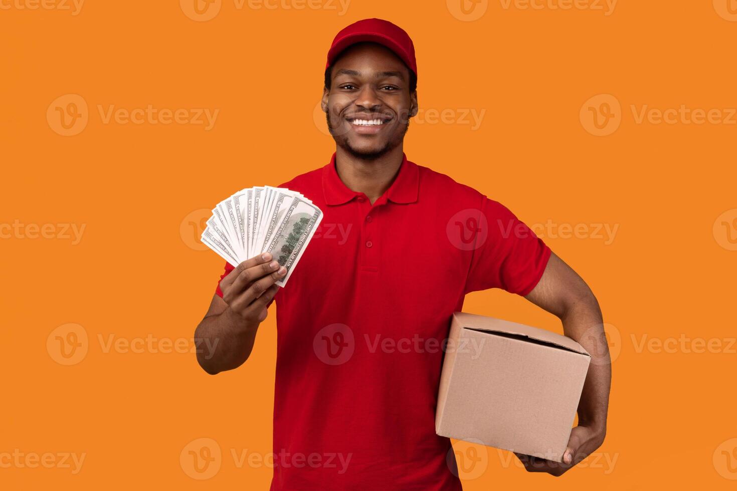 Cash On Delivery. Smiling african american male courier in red cap and uniform holding money and cardboard parcel box, carrying package, standing isolated over orange studio background photo
