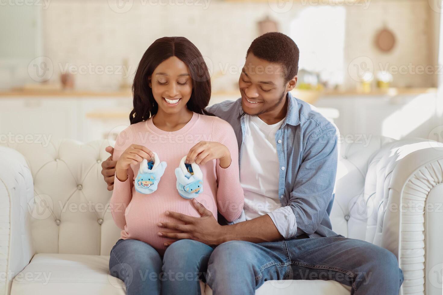 Happy black couple expecting new child, sitting on sofa together and holding baby booties. Caring African American husband tenderly touching his pregnant wife's big belly, expressing his love photo