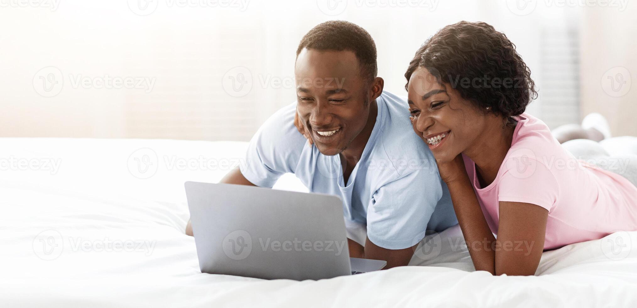 Loving black man and woman husband and wife using modern laptop while relaxing in bed at home, watching photos or websurfing together, enjoying lazy weekend in bedroom, copy space