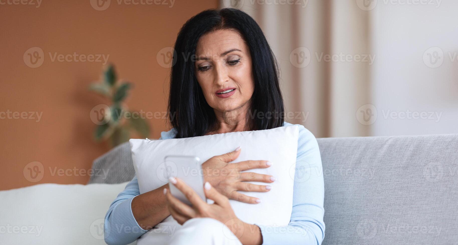 Remote Distant Communication. Senior woman with dark long hair holding mobile phone, making online call and talking to children or watching , embracing pillow, sitting on sofa at home photo