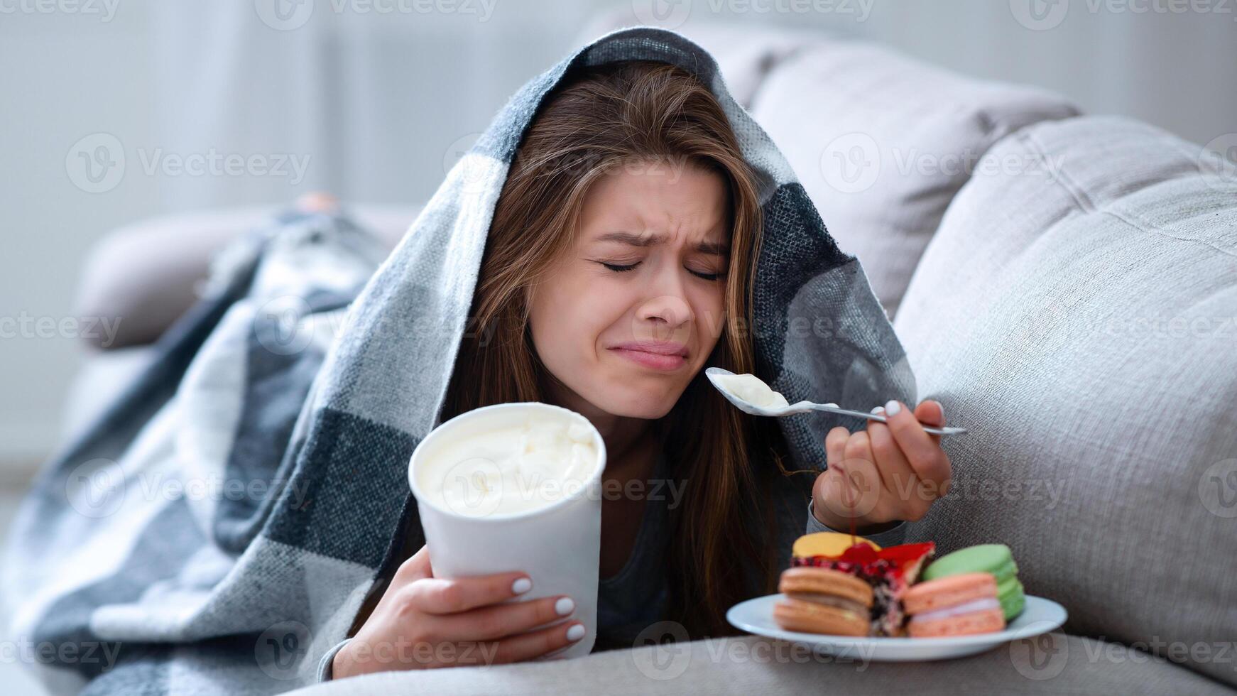 Upset young woman dealing with depression by eating ice cream and sweets under blanket on sofa. Stressed millennial lady coping with personal or relationship problem by comfort food photo