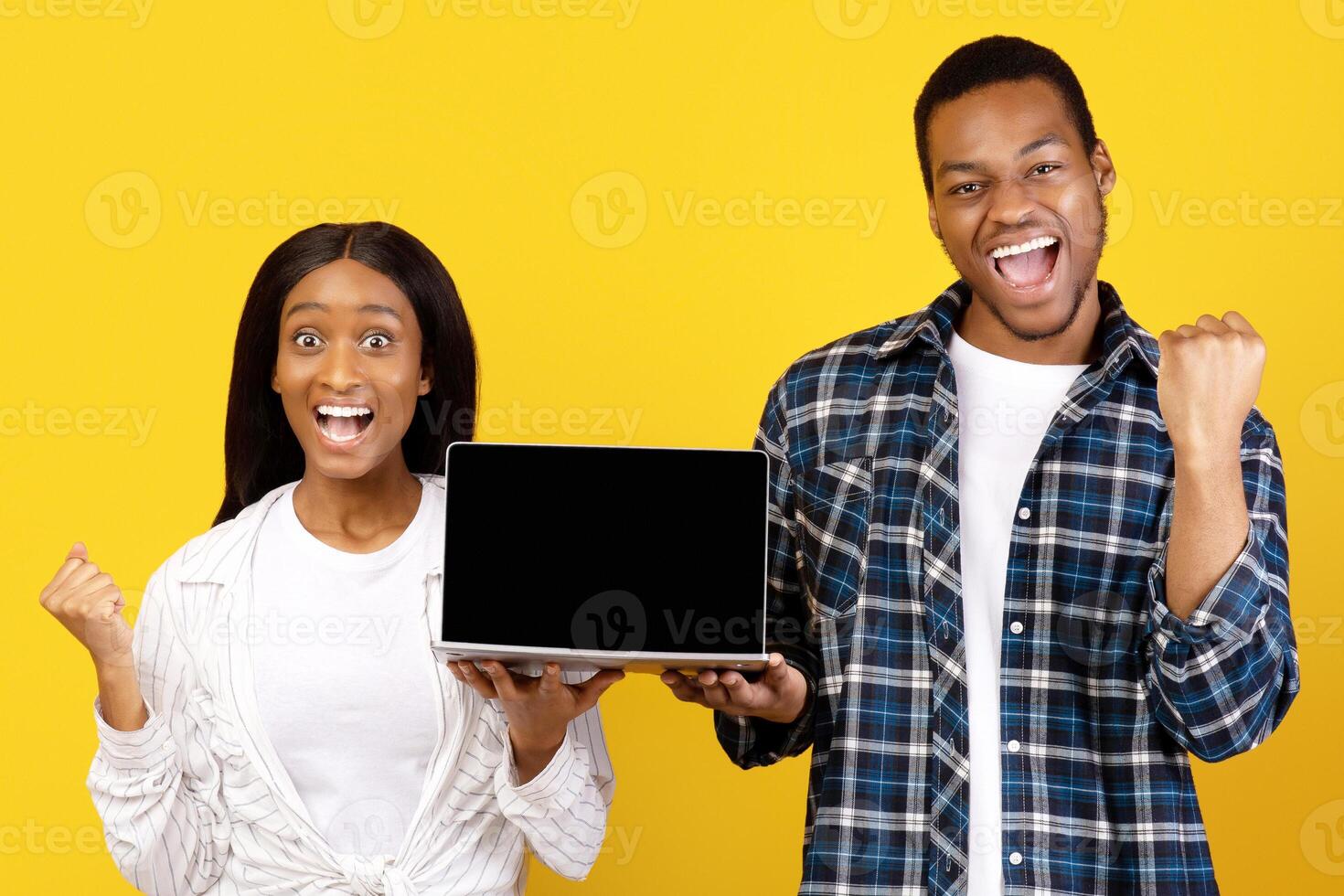 Coveted purchase and online shopping. Happy millennial african american guy and lady expresses emotions of success and keep laptop with empty screen, isolated on yellow background, studio shot photo