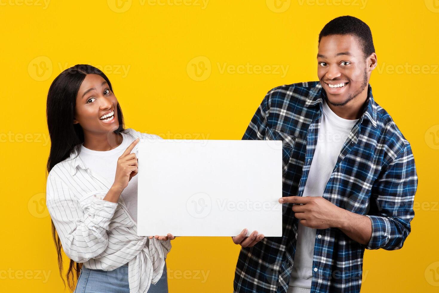 Interesting event, great offer and advice from couple. Cheerful millennial african american family in casual hold banner, pointing fingers to free space, isolated on yellow background, studio shot photo