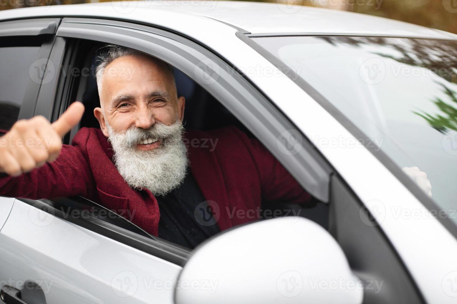 Smiling elderly businessman in stylish suit driving luxury car, looking through car window and showing thumb up at camera, going to office for business meeting. Aged bearded man buying new car photo