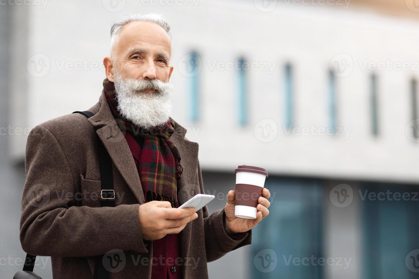 Handsome aged grey-haierd man holding paper cup with coffee and using modern smartphone, walking by street during break, copy space. Elderly man going to job, drinking morning coffee photo