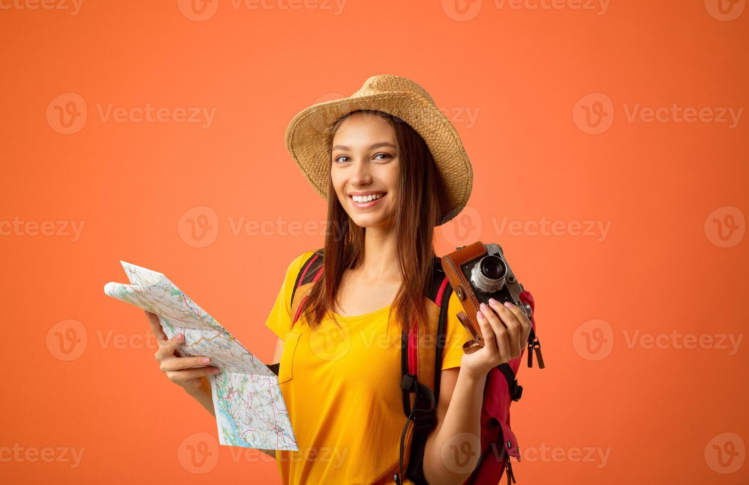 Happy young woman backpacker with camera holding city map over yellow studio background. Joyful lady traveller in summer hat looking at camera and smiling, going for sightseeing abroad, copy space photo