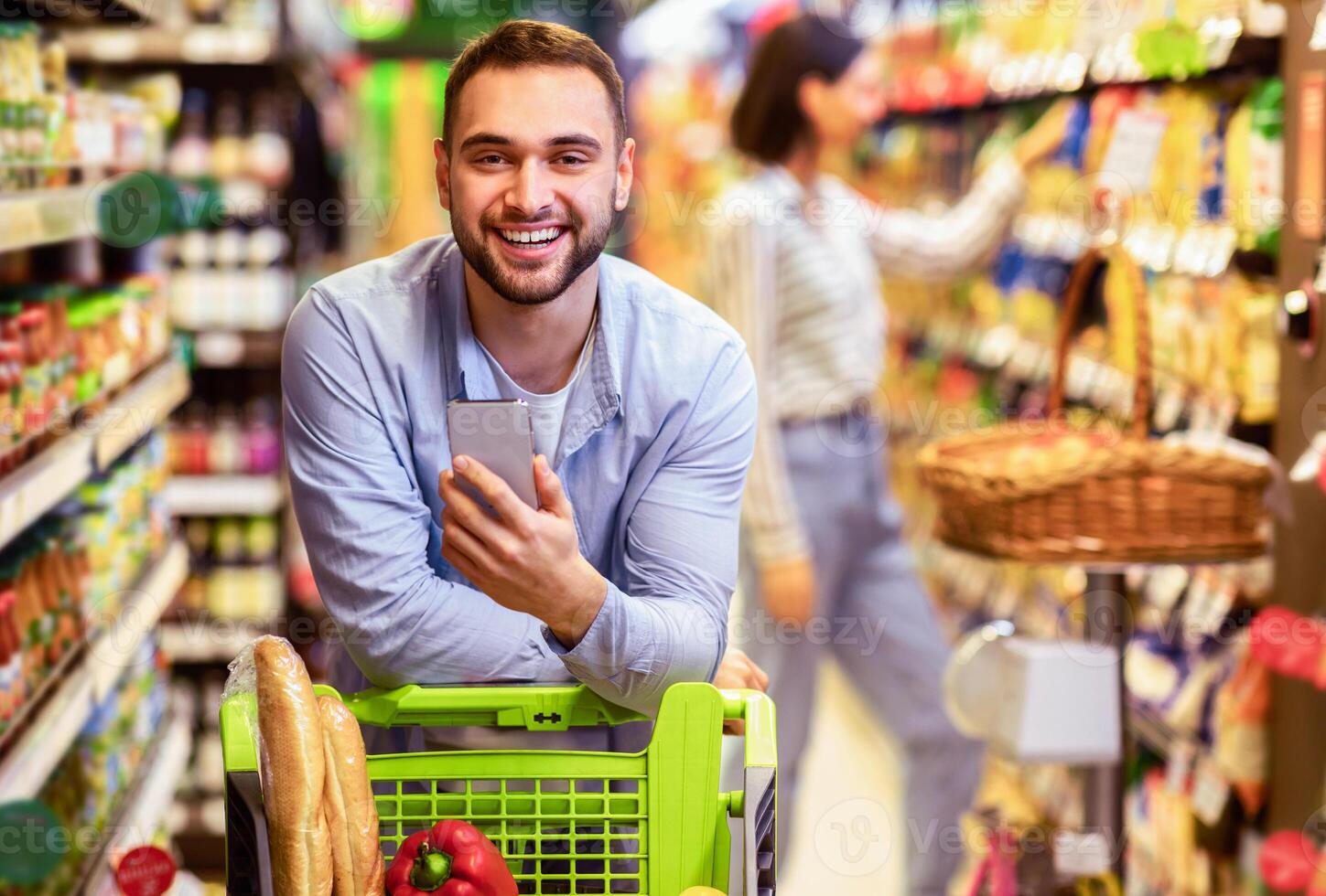 Portrait of smiling young casual man leaning on shopping trolley cart indoors, using his smartphone, posing and looking at camera. Satisfied male customer buying products in local grocery store photo