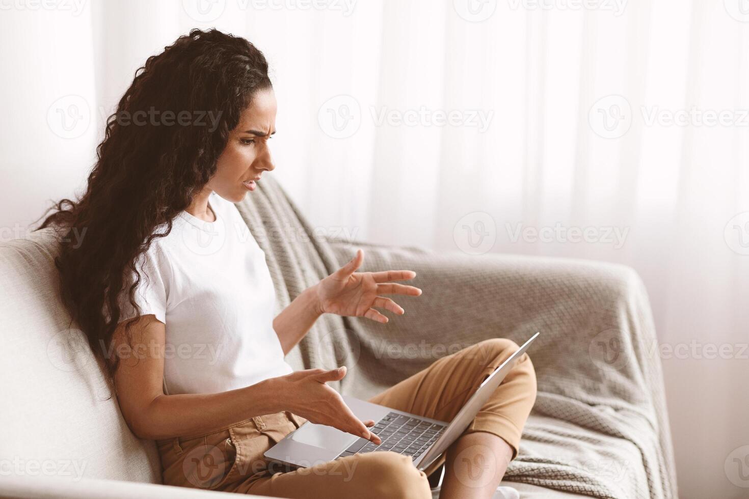 Side view of annoyed young woman looking at laptop screen and gesturing, copy space. Angry millennial lady sitting with computer on couch in living room, working online, having difficulties photo
