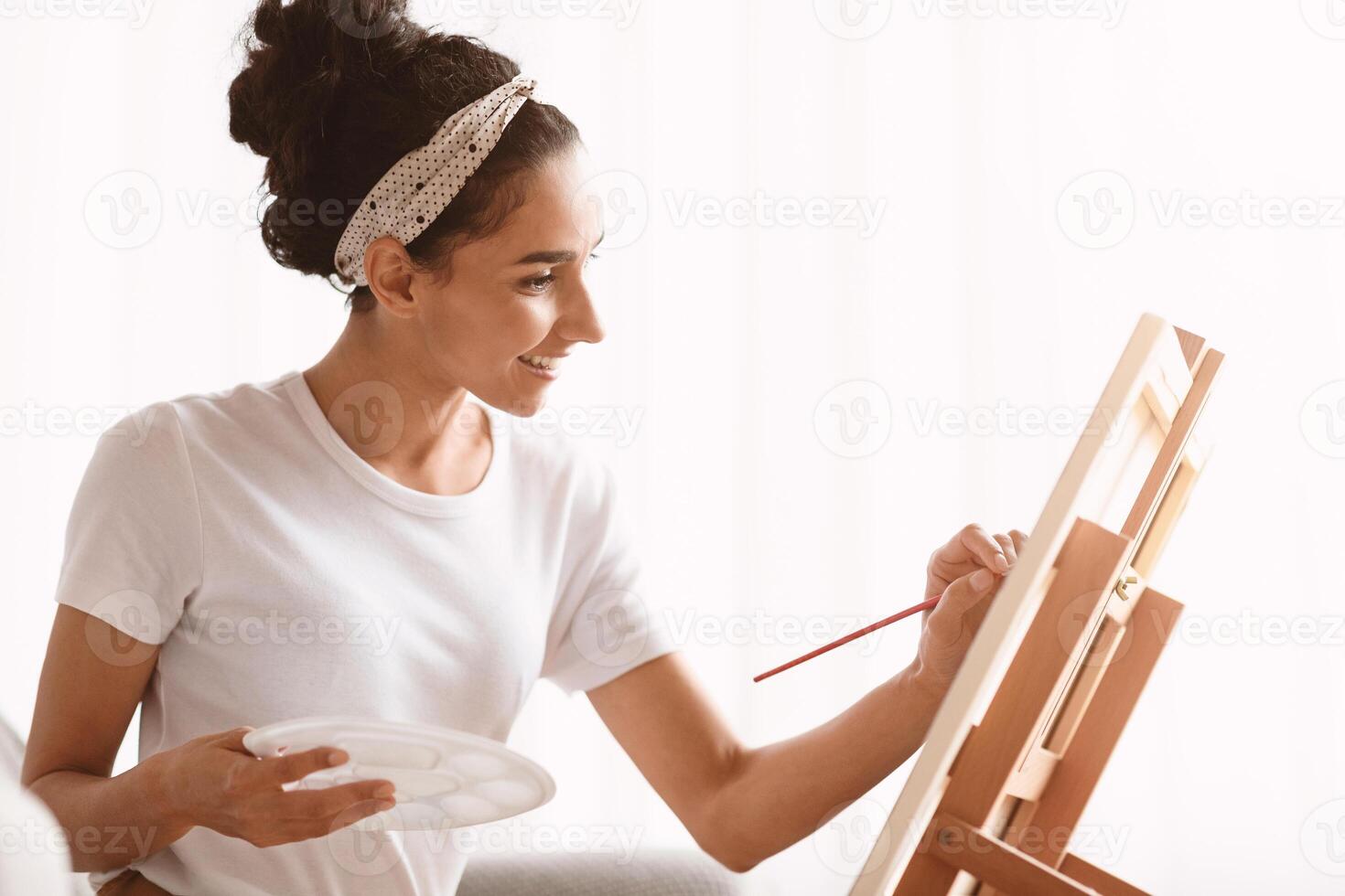 Closeup of joyful brunette woman sitting on sofa and painting, using palette, brush and easel. Creative lady enjoying new hobby, painting still life at home, copy space. Domestic lifestyle concept photo