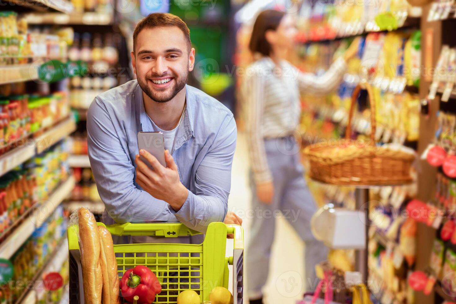 Couple In The Supermarket. Portrait of smiling young bearded man leaning on shopping trolley cart indoors, using mobile phone, his girlfriend is choosing food in the blurred background photo