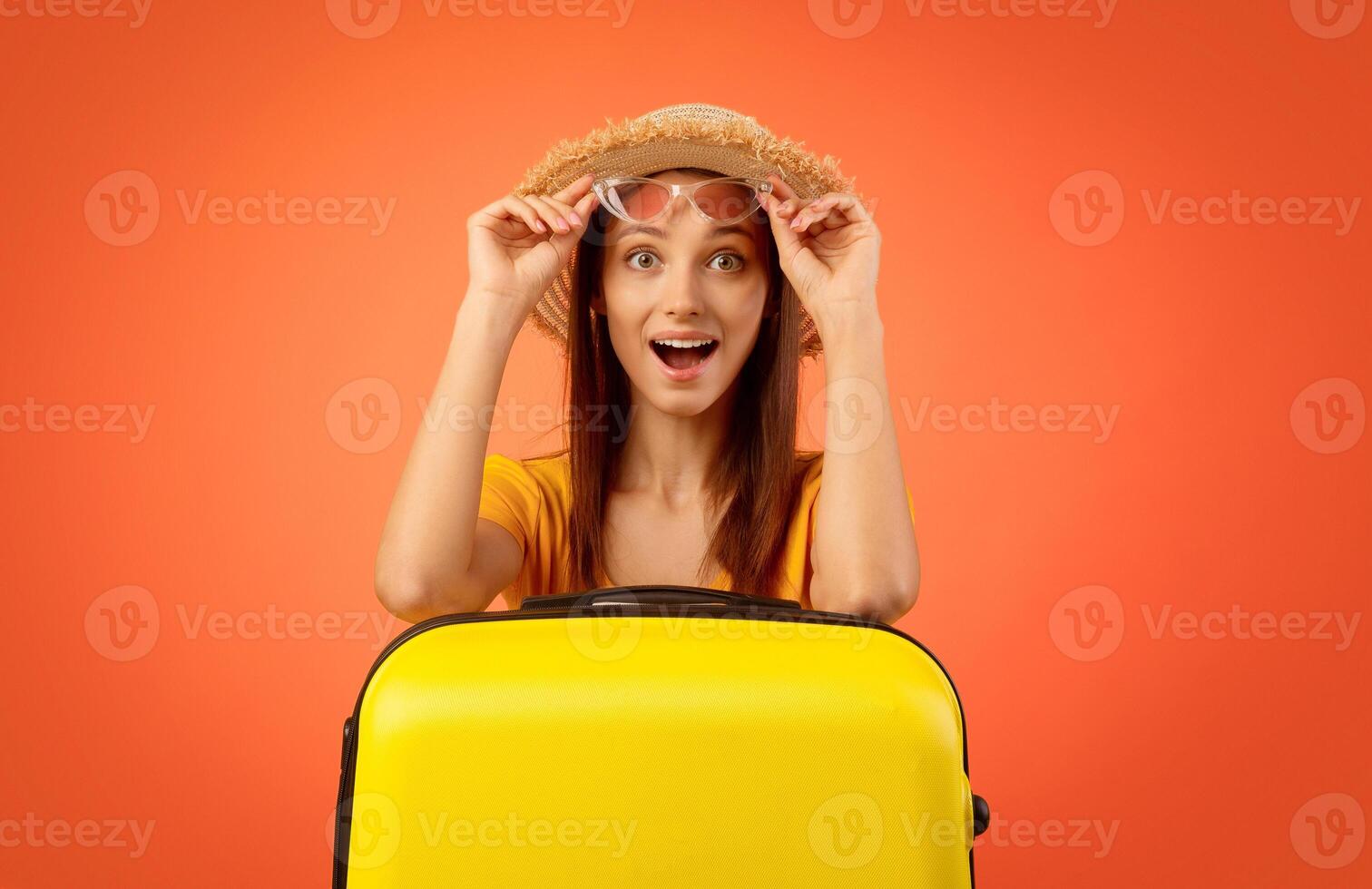 Amazed girl holding sunglasses and leaning on suitcase, yellow studio background, time to travel photo
