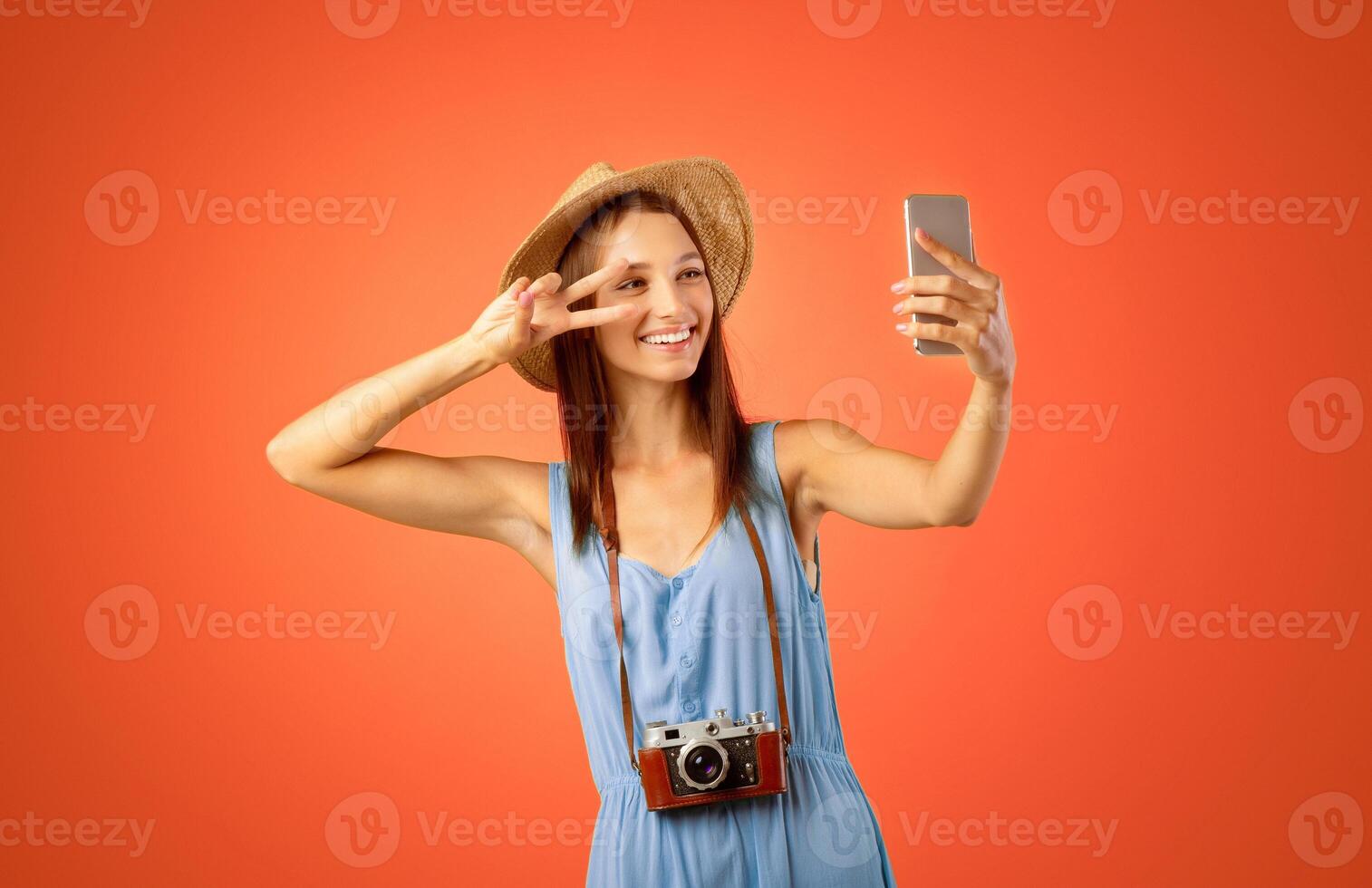 Pretty young lady traveller taking selfie on mobile phone, yellow studio background. Joyful woman with camera in summer hat taking photo of herself for social media, using smartphone, gesturing
