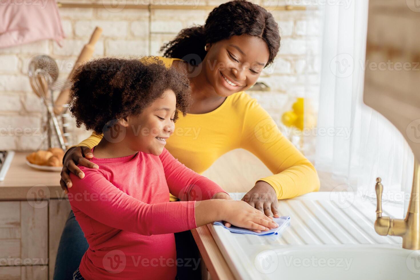Loving Black Mother Teaching Her Daughter Housekeeping, Cleaning Kitchen Together. Cute Little Girl Wiping Table With Cloth And Smiling, Enjoying Helping With Household Chores, Free Space photo