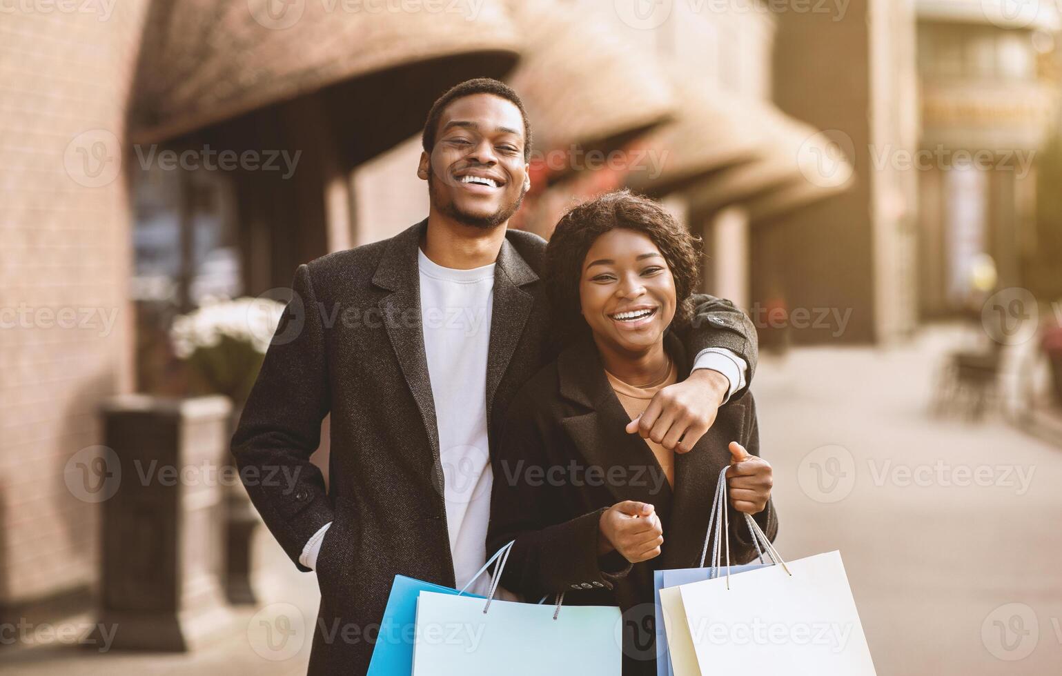 Shopaholics after walking in mall during Black Friday and seasonal sale before holidays. Happy african american husband and wife in coats hold many colored bags and hug, outdoors, copy space photo