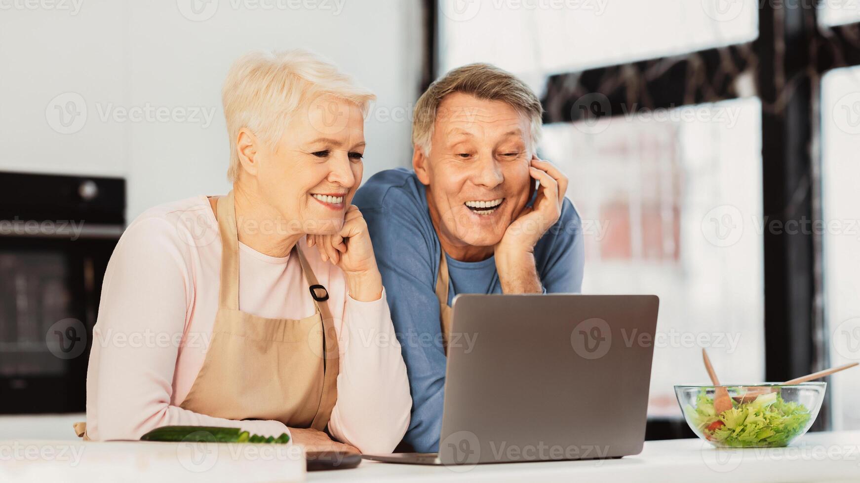 Senior Spouses Calling Sitting At Laptop In Modern Kitchen At Home. Happy Retired Lifestyle. Elderly Couple Cooking And Browsing Internet On Computer Together. photo