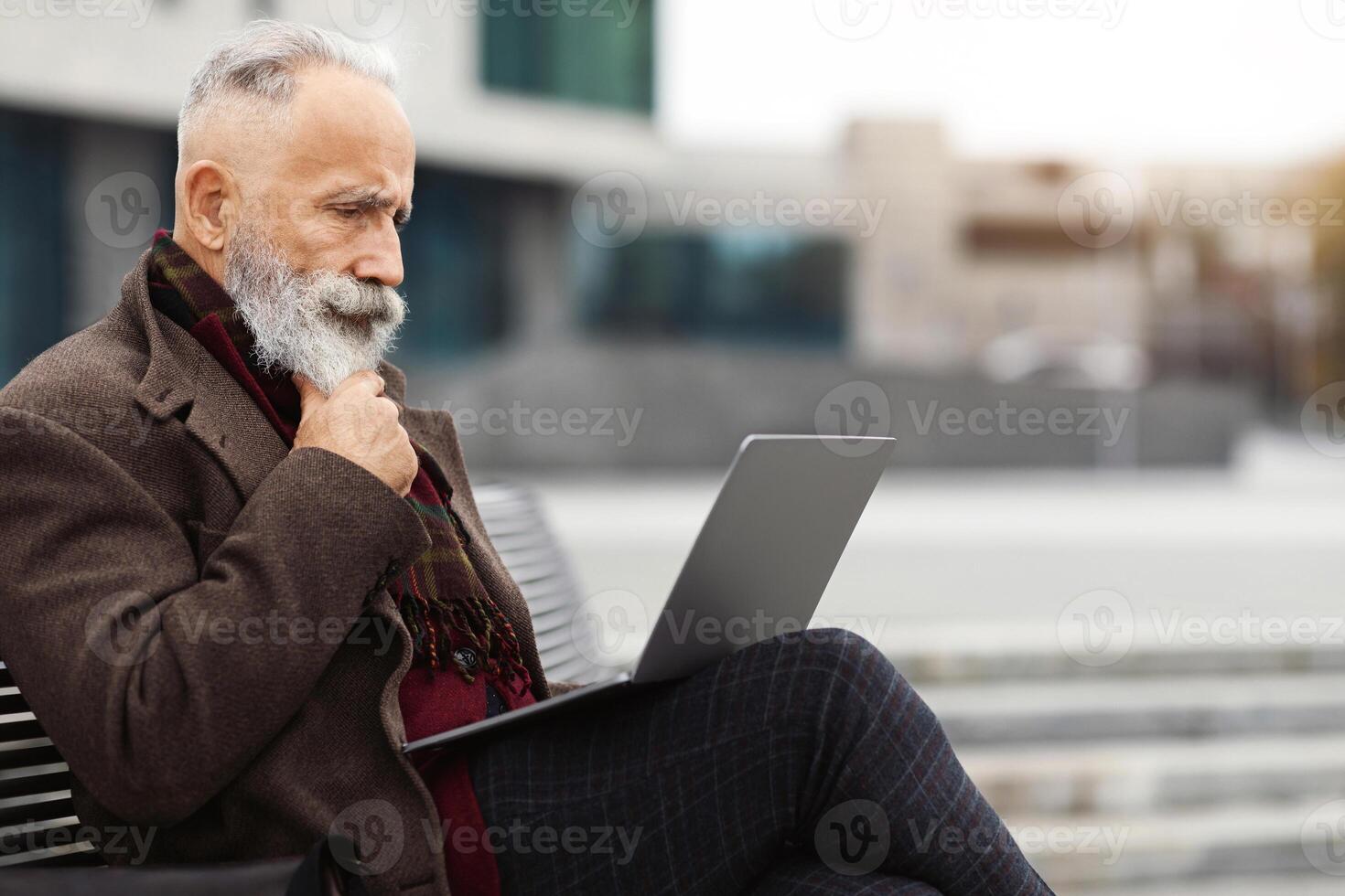 Pensive senior bearded businessman with laptop sitting on bench, typing on keyboard, sending email, empty space. Stylish aged grey-haired man sitting on street by business center, working online photo