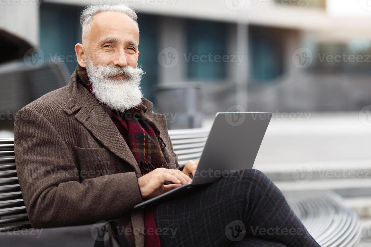Wealthy grey-haired aged man with long beard in warm stylish outfit sitting on the bench by office building, using laptop, working online, chatting with clients or websurfing, empty space photo