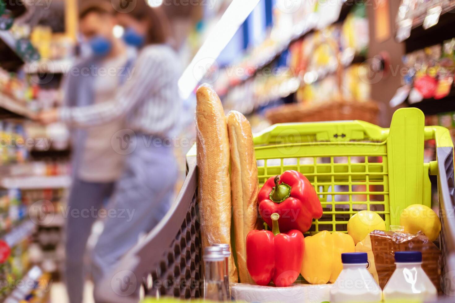 Family Shopping And Healthy Products. Young Couple Standing In Supermarket In The Blurred Background, Selective Focus On Shopping Trolley Cart Full Of Food, Vegetables, Bread And Dairy photo