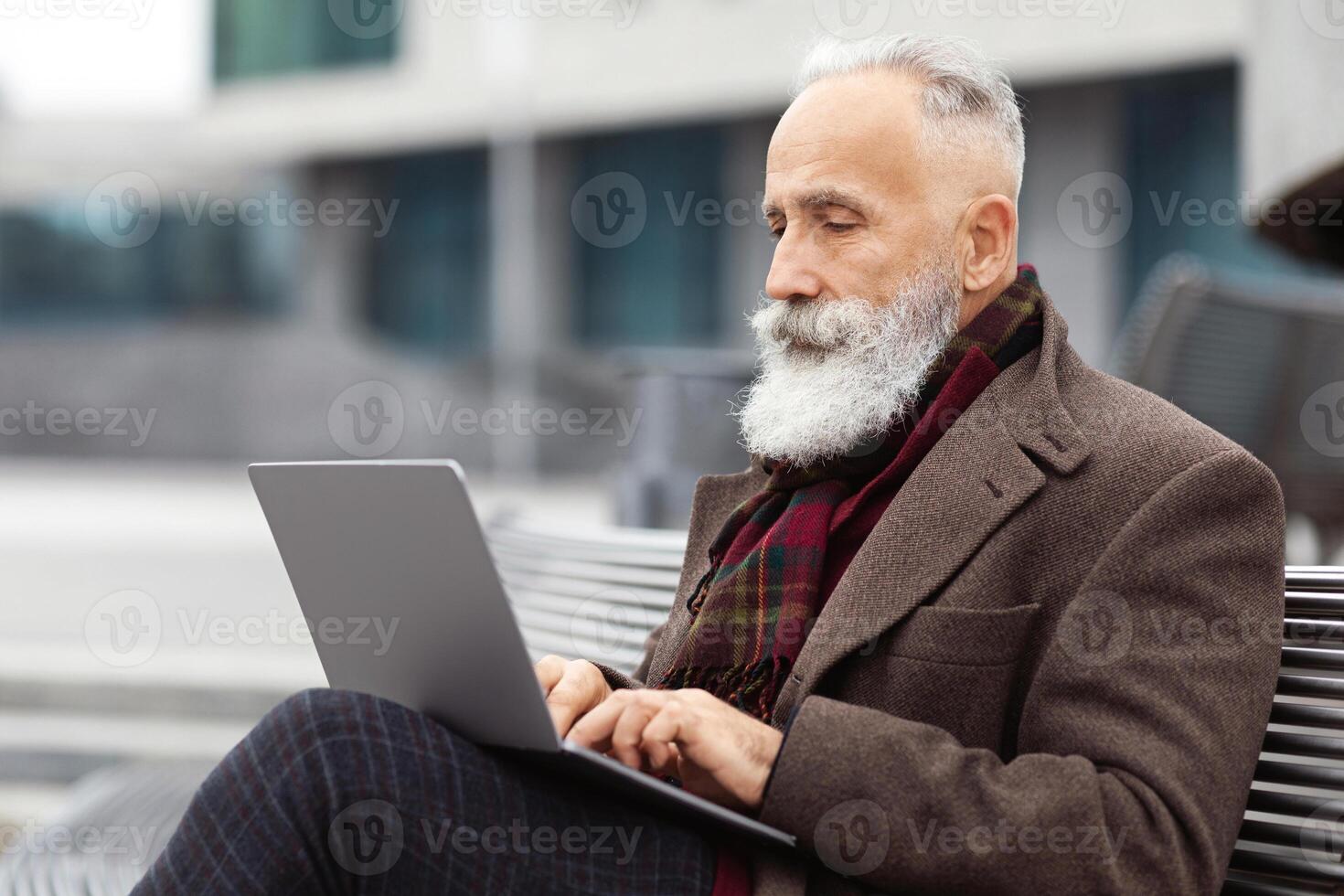 Concentrated grey-haired aged man with long beard in warm stylish outfit sitting on the bench by office building, using laptop, working online, chatting with clients or websurfing, empty space photo