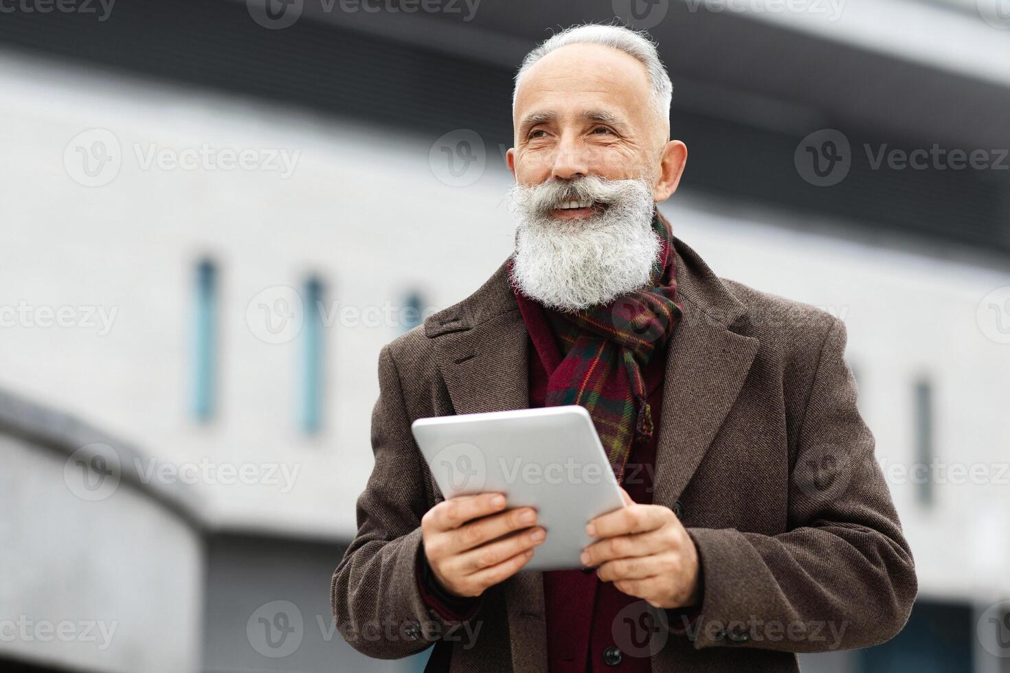 Cheerful aged man with long grey beard holding digital tablet while walking by street, looking at copy space and smiling. Senior businessman in stylish outfit with pad standing by office building photo