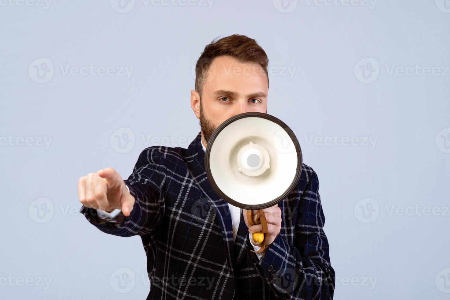 Determined young man in formal wear shouting into megaphone and pointing at you on grey studio background. Public speaker with bullhorn making announcement, proclaiming message photo