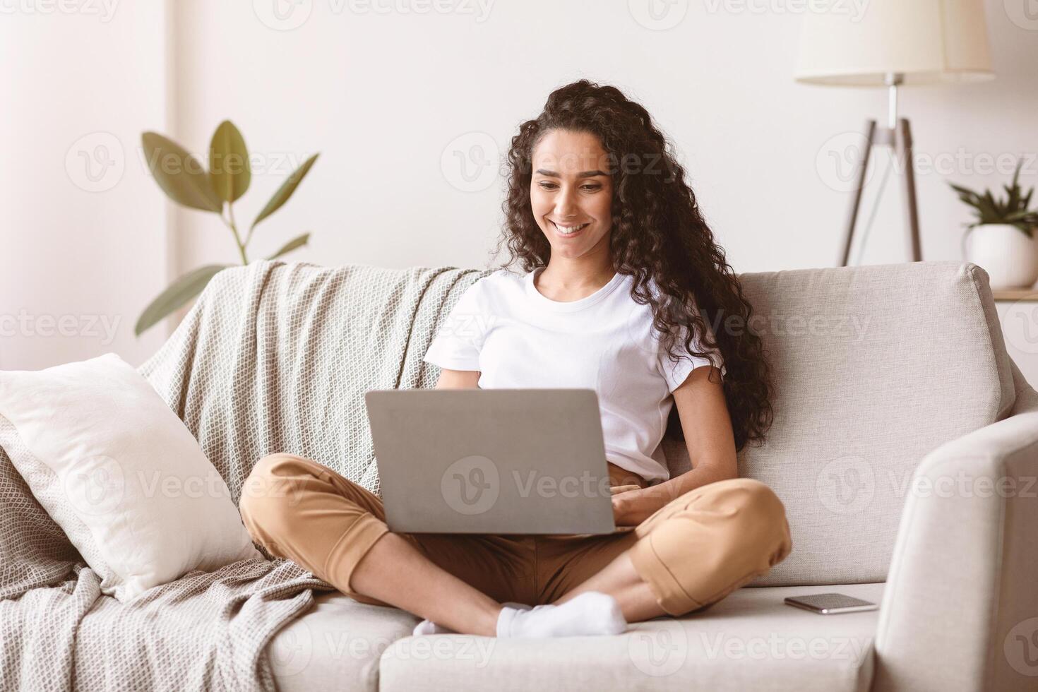 Happy young woman with dark curly hair freelancer working from home, sitting on couch and using modern laptop, typing text or communicating with clients online, blogging or copywriting, copy space photo