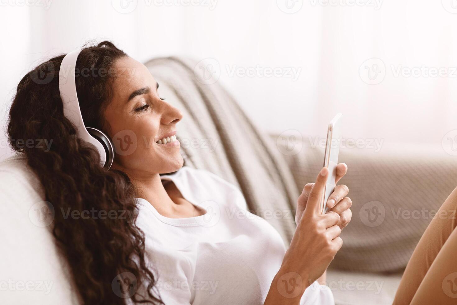 Happy young brunette woman with long curly hair in headphones using mobile phone, watching movie or listening to music, using mobile application, sitting on couch at home, copy space, side view photo