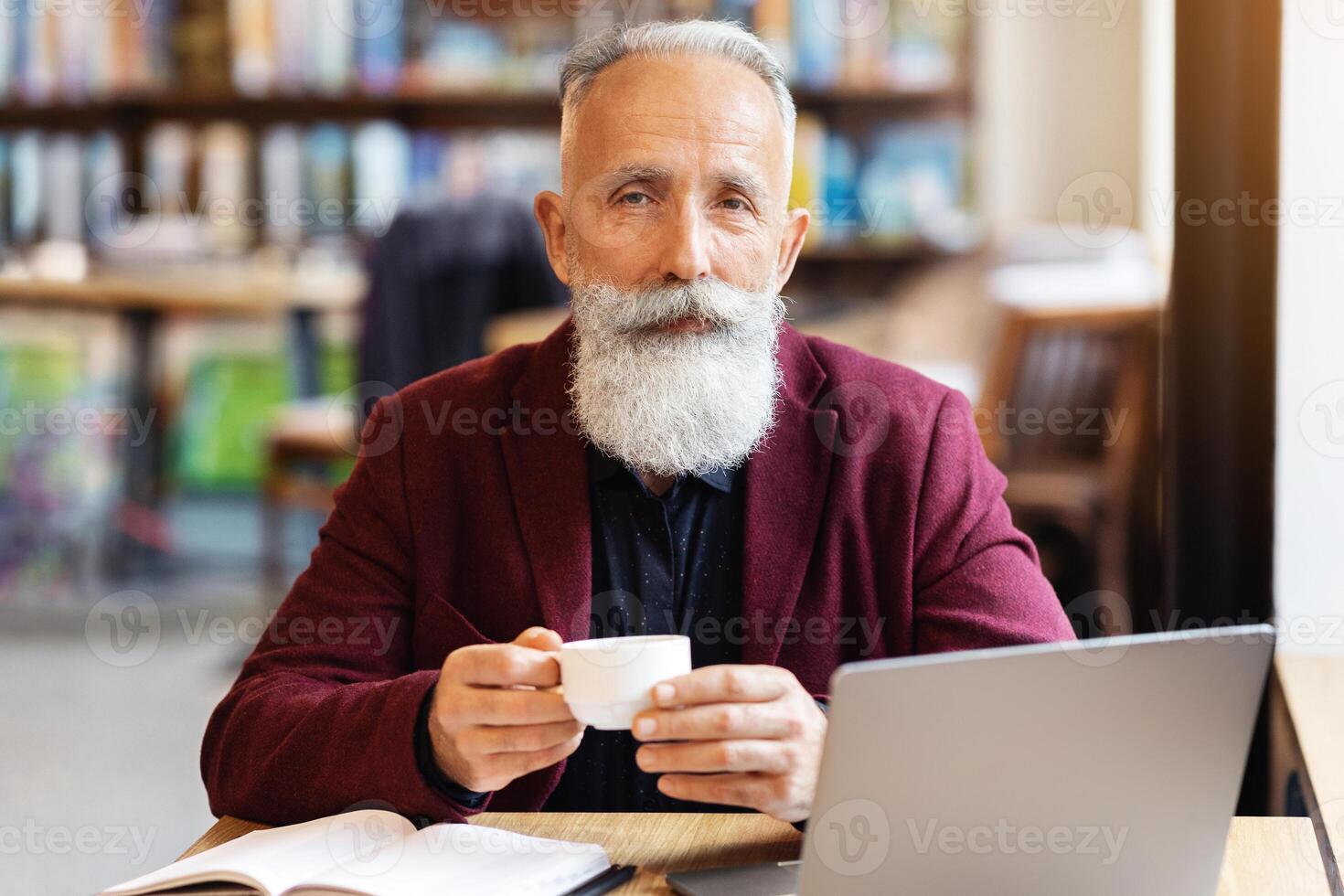 Handsome elderly man with long grey beard planning his day while having morning coffee at cafe, sitting at desk with laptop and notebook, copy space. Senior businessman taking notes and working online photo