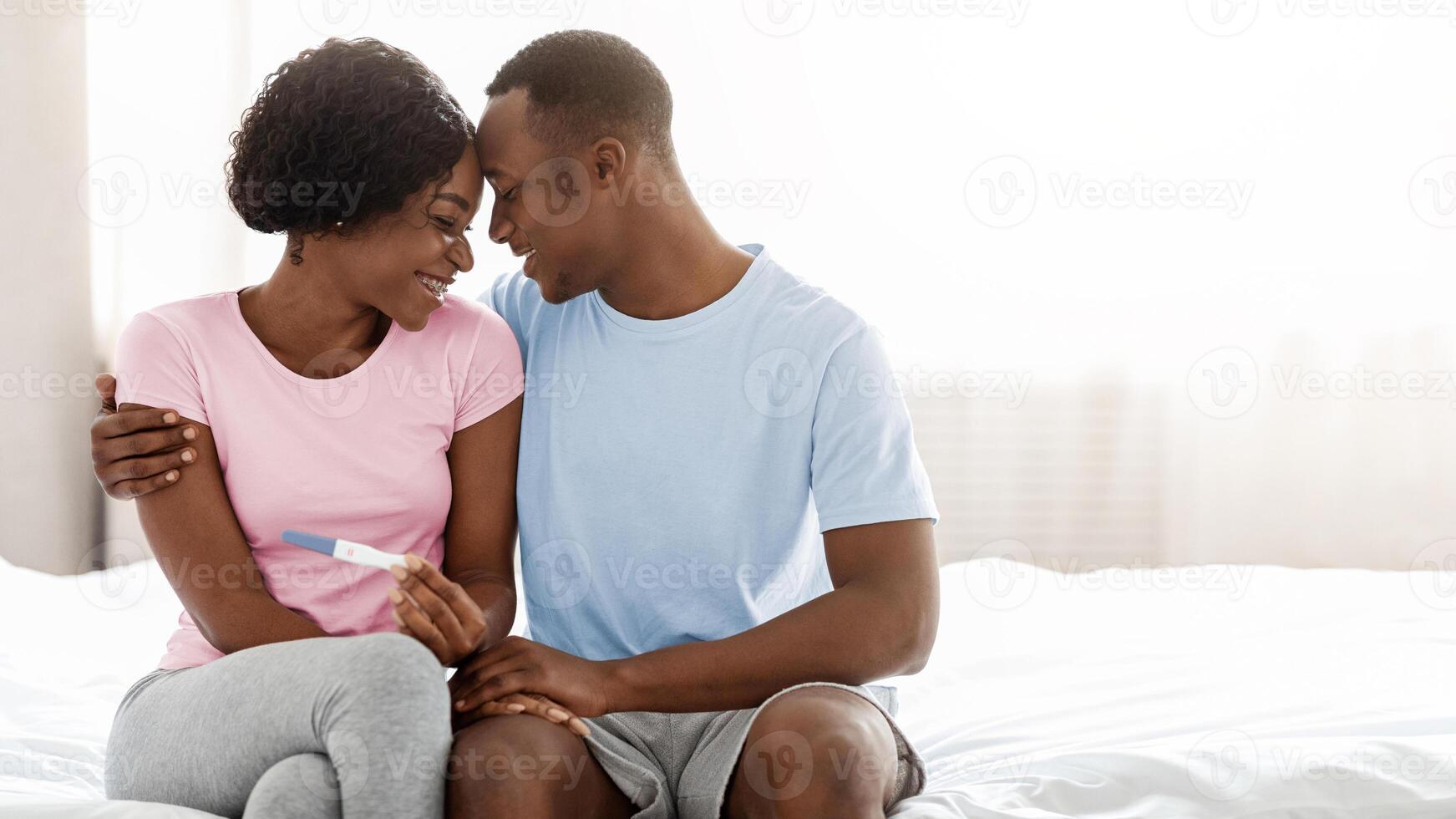Young black couple will become parents, holding positive pregnancy test and touching each other with foreheads, bedroom interior, empty space. Delighted african american family waiting for baby photo