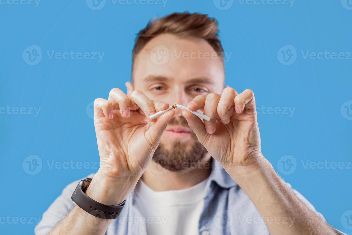 Stop smoking concept. Young bearded guy breaking cigarette on blue studio background, selective focus. Millennial man fighting his addiction to nicotine, saying NO to harmful habit photo