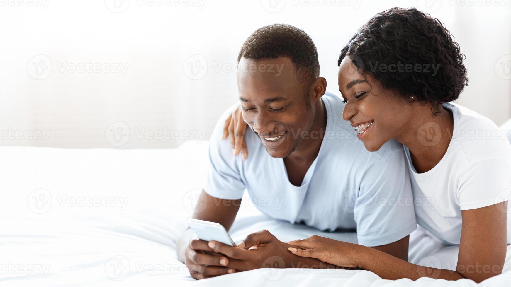 Joyful black couple holding mobile phone, using new application together, lying on bed at home, copy space. African american young man and woman using smartphone in bedroom, having fun together photo