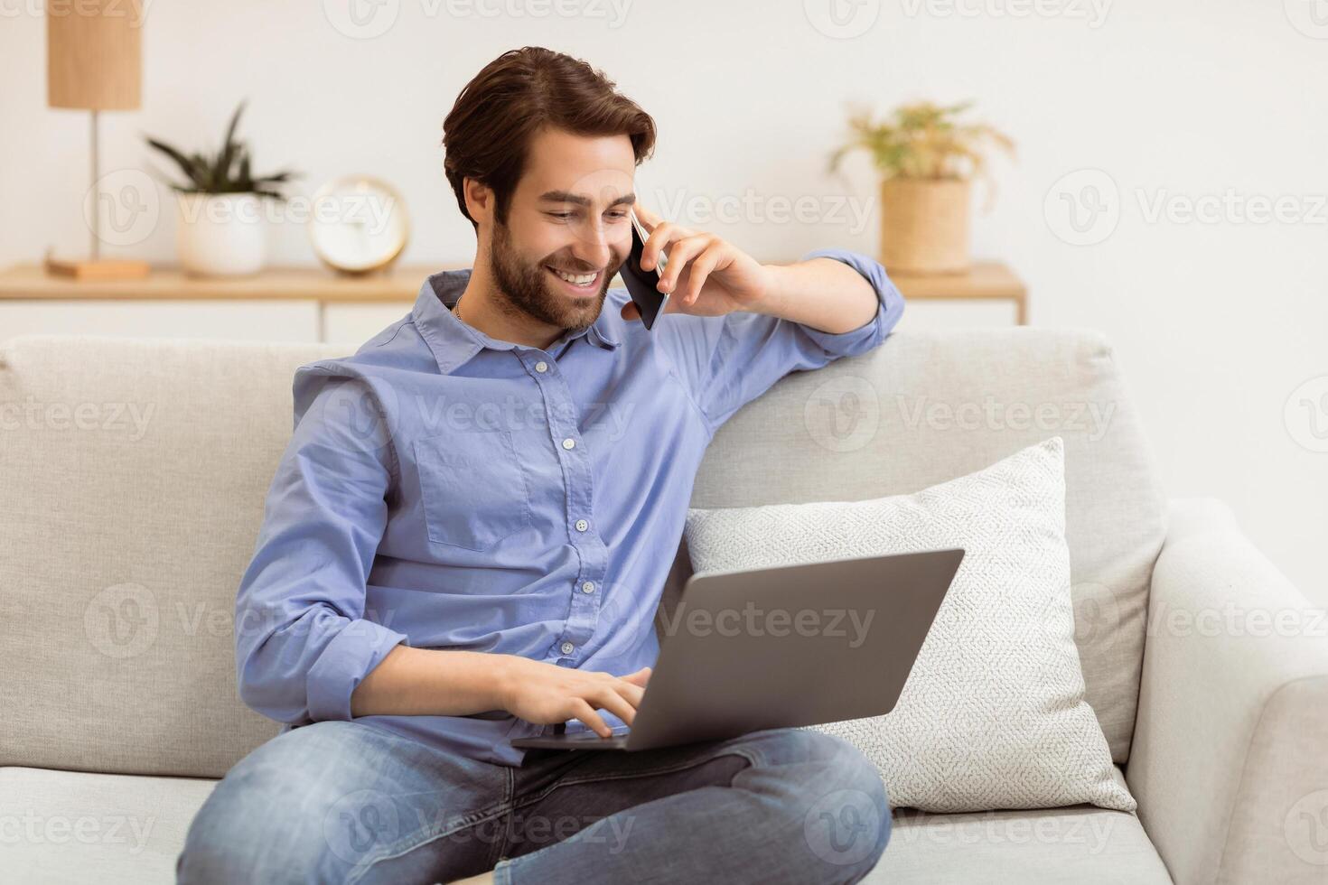 Happy Man Talking On Cellphone At Laptop Computer Sitting On Couch In Modern Office. Freelancer Guy Speaking By Phone Working Indoors. Freelance, Mobile Communication Concept photo