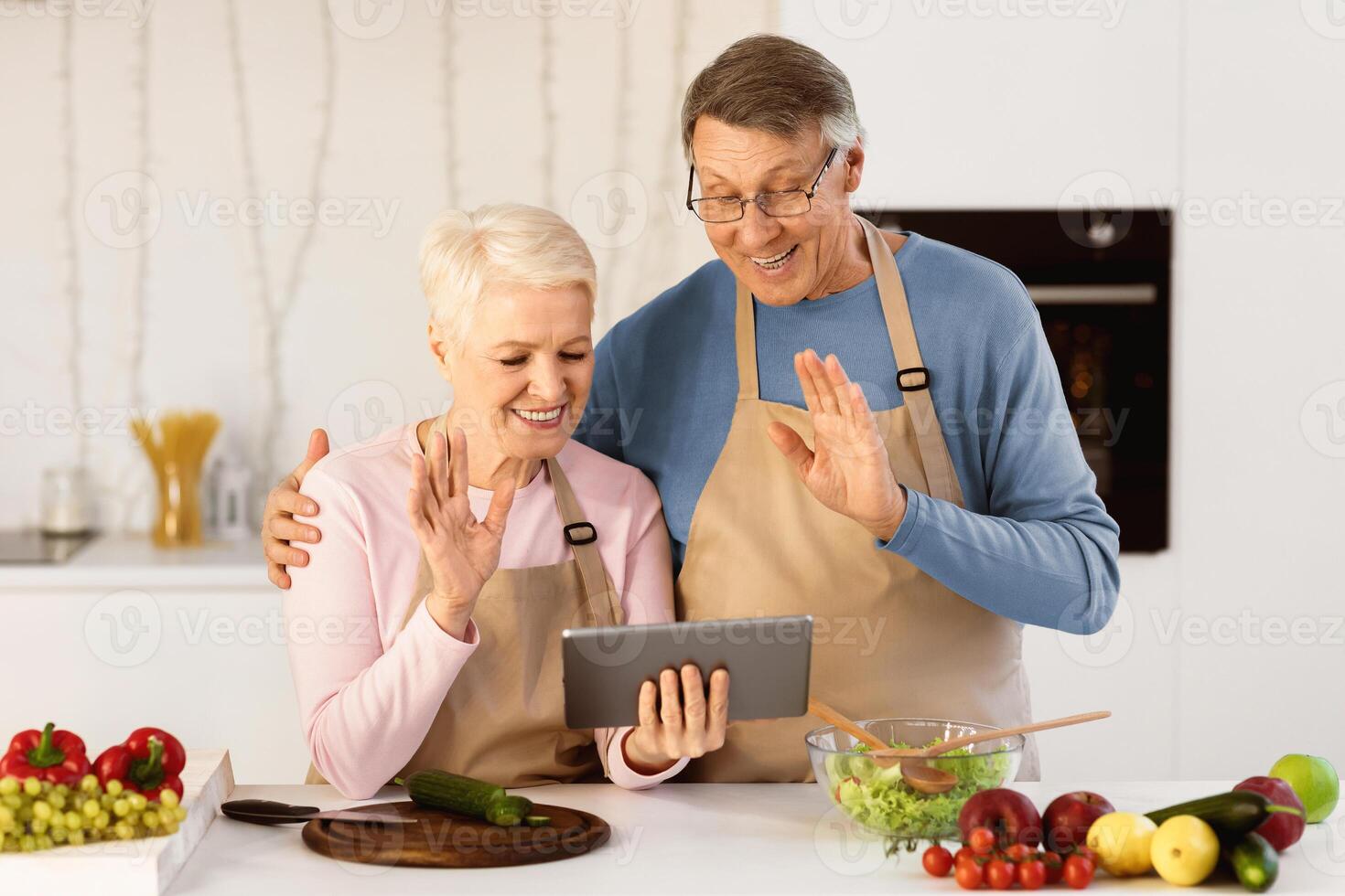 Happy Senior Couple Calling Using Digital Tablet, Waving Hand To Computer Webcam Standing In Modern Kitchen Cooking Together At Home. Virtual Remote Communication By Call Concept photo