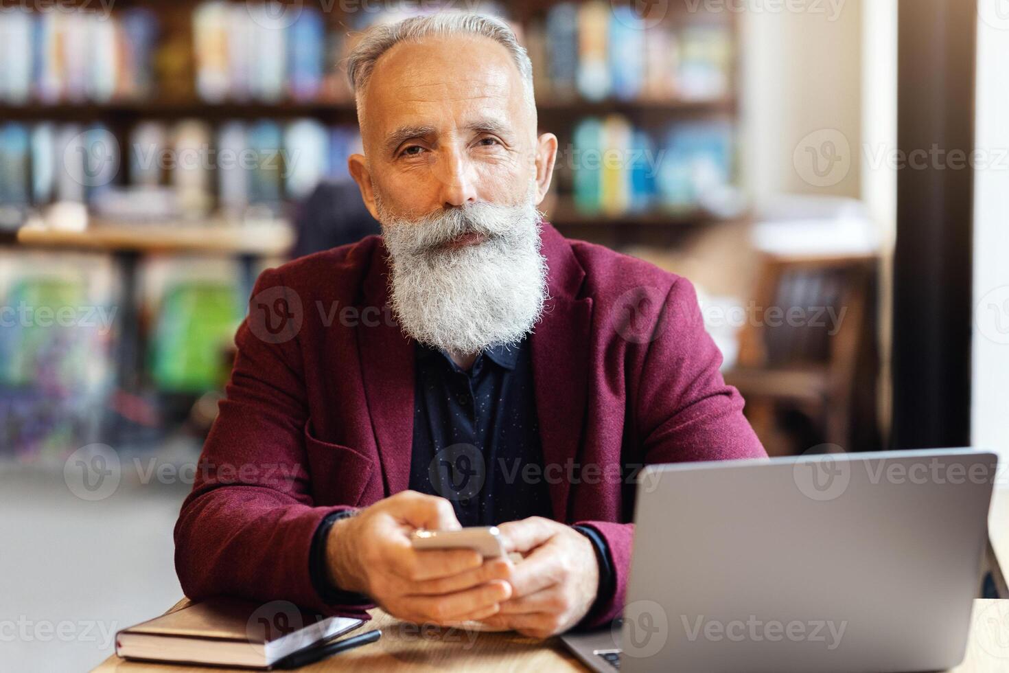 Wealthy elderly man using smartphone at cafe, sitting in front of laptop, freelancing or sending emails during lunch break. Aged businessman working online at cafe, using computer and mobile phone photo