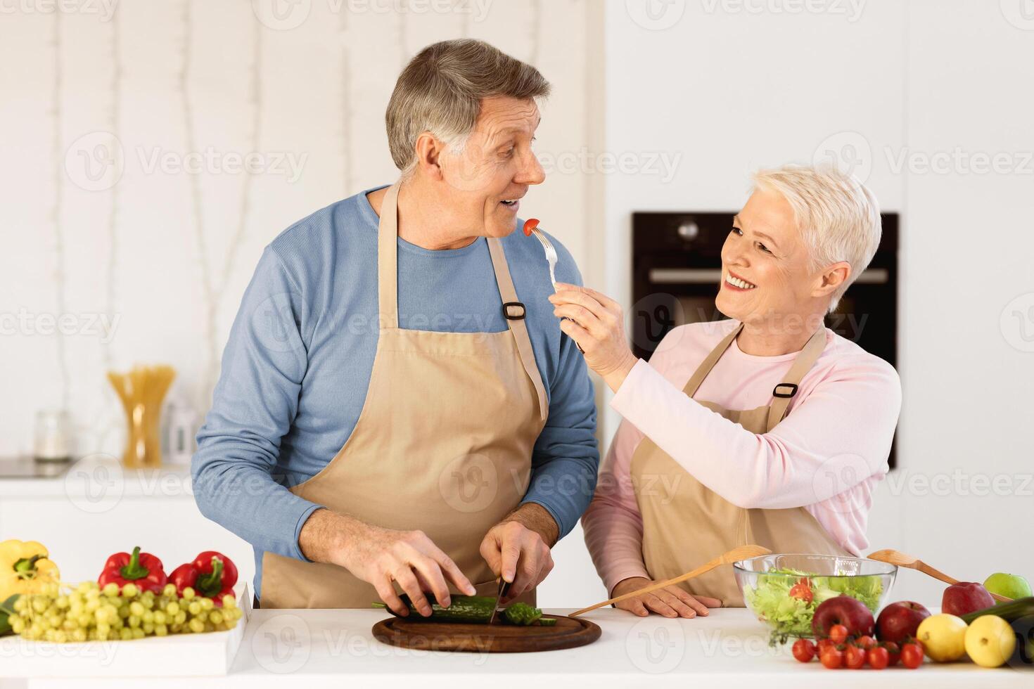Aged Spouses Cooking And Tasting Food Feeding Each Other Having Fun During Dinner Preparation Standing In Modern Kitchen At Home. Senior Family Couple Enjoying Weekend Preparing Meal Together. photo