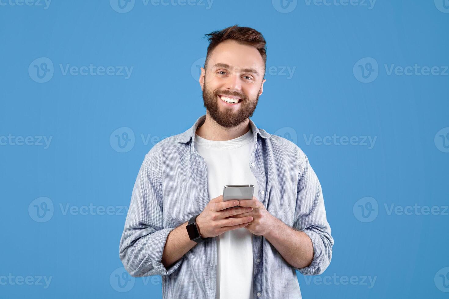 Portrait of cool bearded guy using mobile phone on blue studio background. Cheerful young man with modern gadget browsing internet, checking email, posting in social media photo