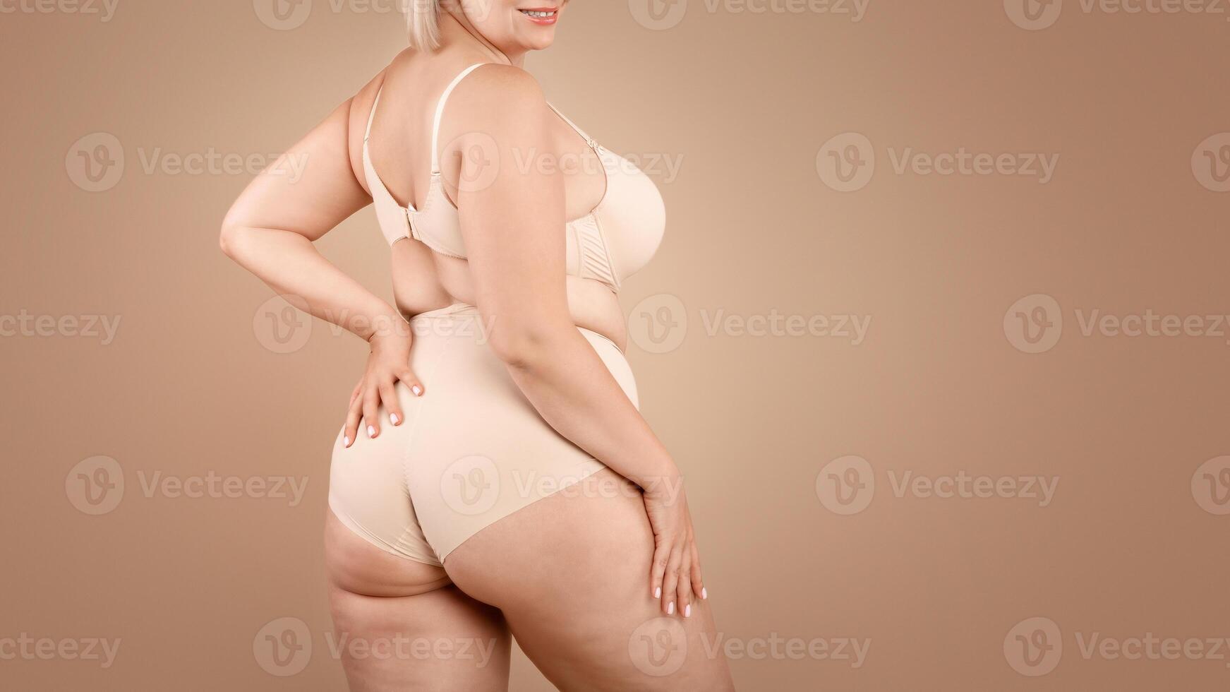 Young plus size woman in beige underwear posing over brown studio background, half-turned at camera, cropped image with free space photo