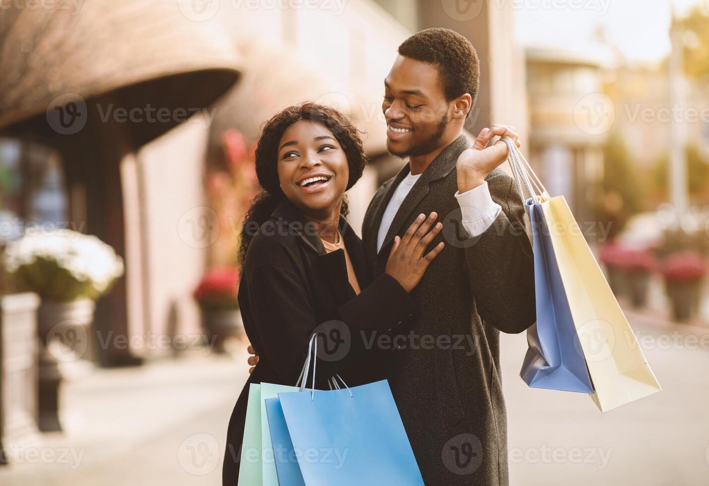Best sale before Halloween, Xmas and New Year holidays. Happy young african american guy and lady in coats hold many colorful packages, hug and express success on street, near store or boutique photo