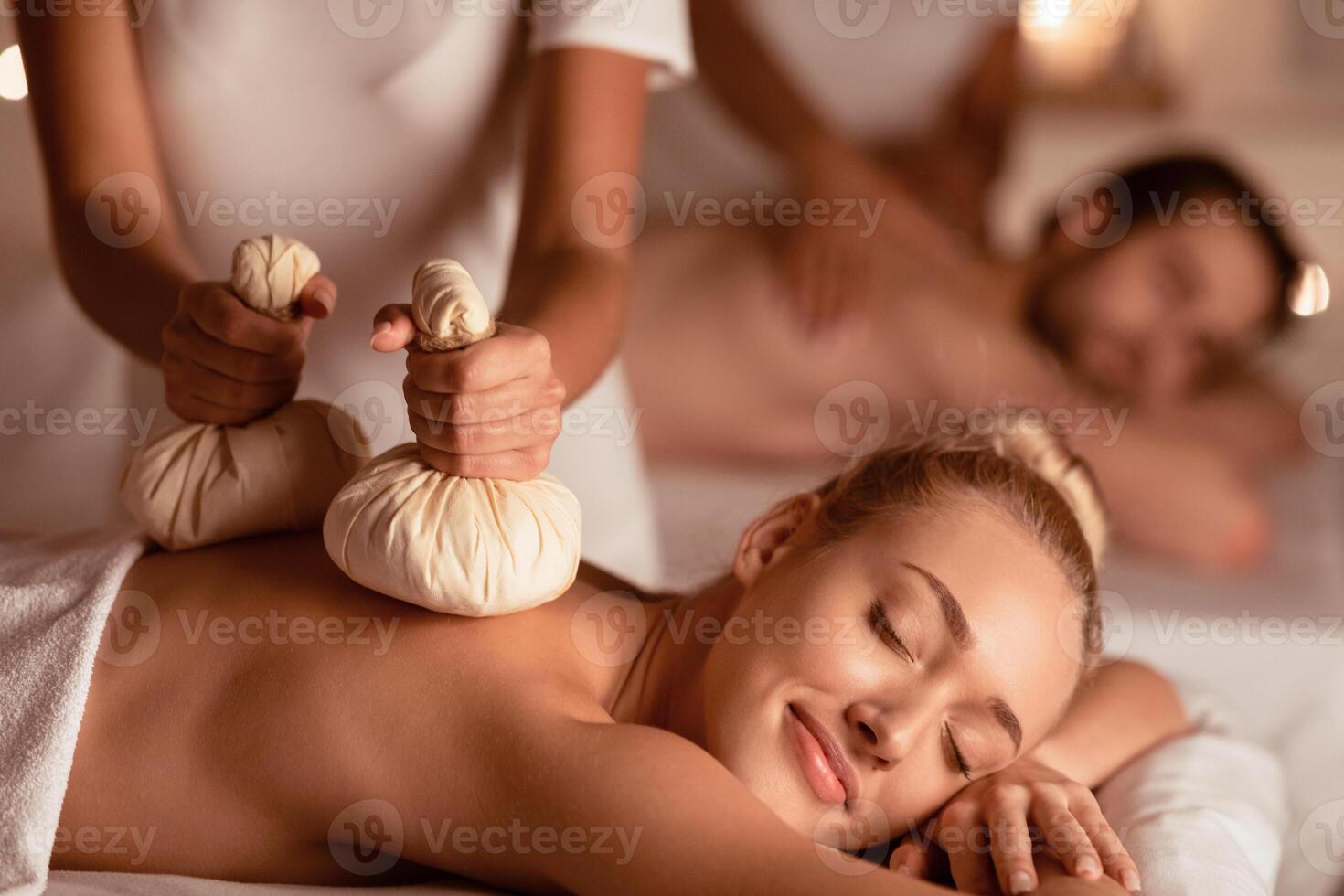 Woman Enjoying Exotic Herbal Massage With Aromatic Bags Herbs During Couple Beauty Treatment At Spa Center Having Romantic Date Or Honeymoon Vacation, Lying With Eyes Closed Indoor. Selective Focus photo