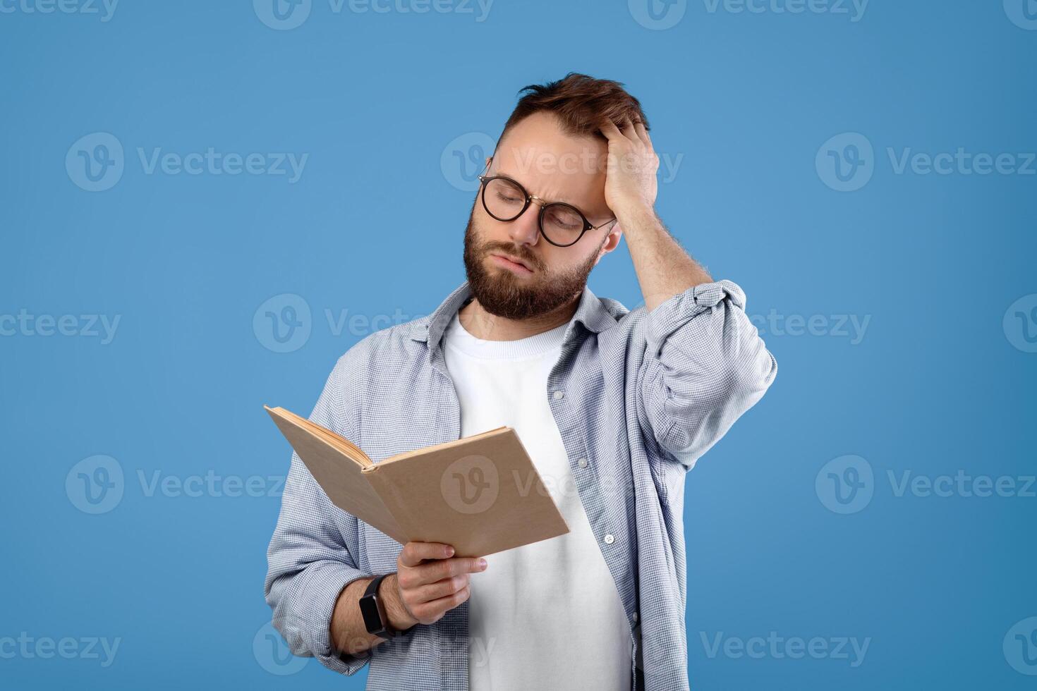 Young Caucasian student having difficulty understanding textbook, studying for exam on blue studio background. Puzzled millennial guy scratching head while reading complicated book photo