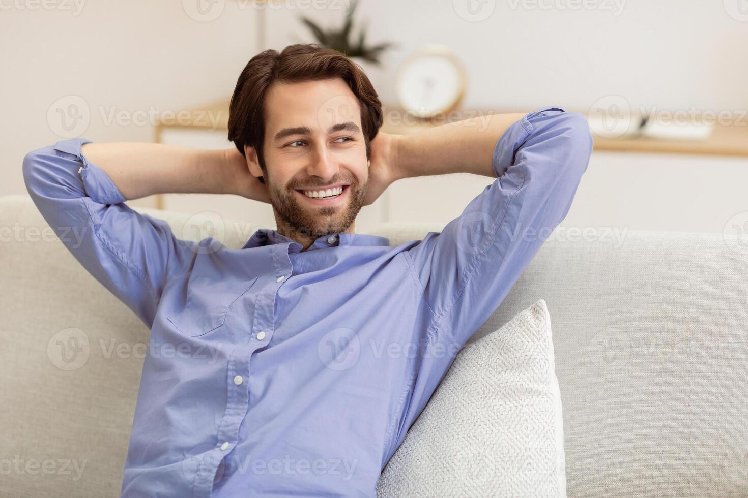 Smiling Man Relaxing Sitting On Couch At Home, Holding Hands Behind Head. Successful Millennial Guy Resting On Sofa Enjoying Comfort And Homey Weekend. Relaxation, Domestic Lifestyle Concept photo