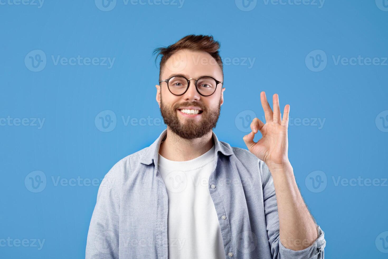 Everything is okay. Portrait of happy Caucasian guy showing OK gesture over blue studio background. Confident young man expressing approval, positivity or support, pleased with something photo