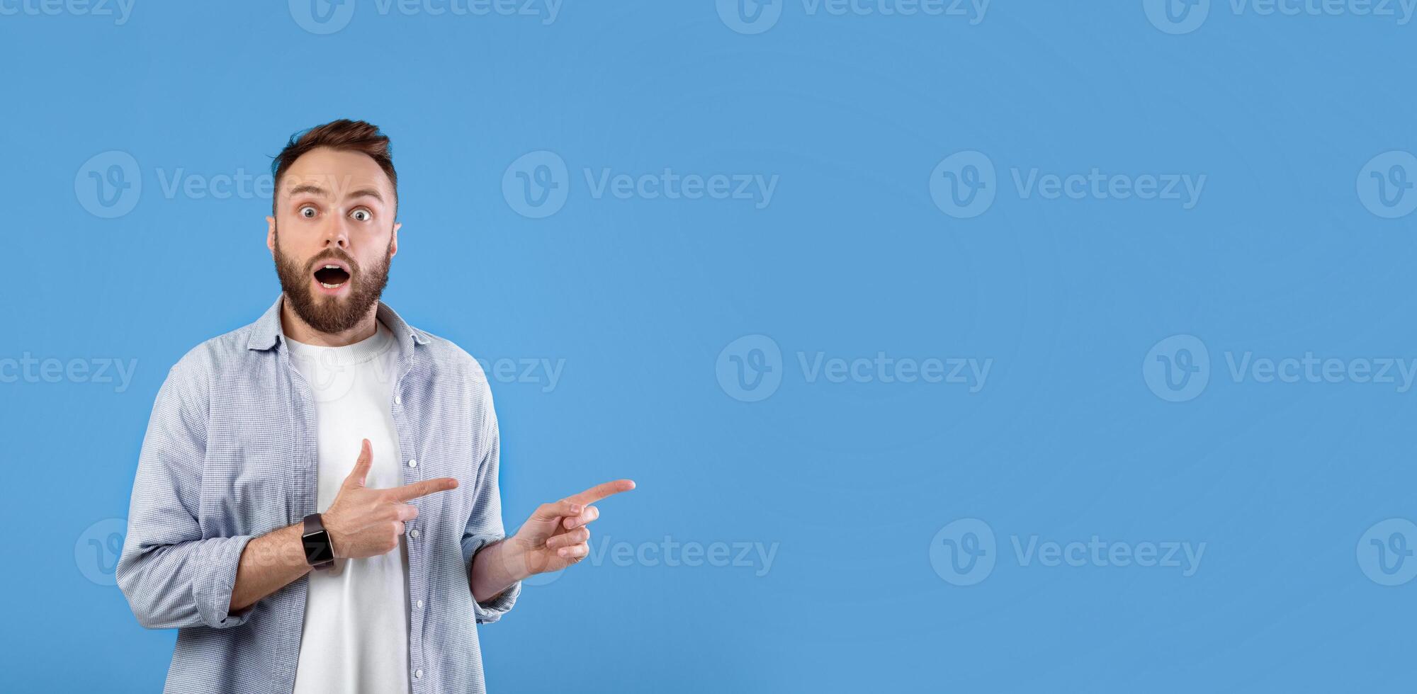 Shocked millennial man pointing at empty space in disbelief, standing with open mouth over blue studio background, free space. Stunned young guy advertising something, offering sale or promo photo