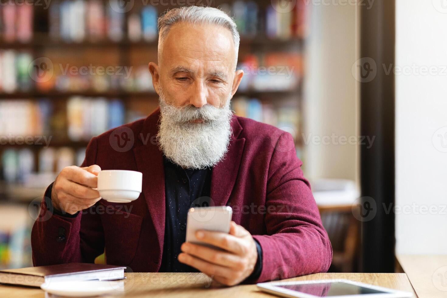 Grey-haired elderly man in burgundy jacket drinking coffee and using mobile phone, checking his email or chatting with clients, resting at cafe alone, empty space. Senior people and gadgets concept photo