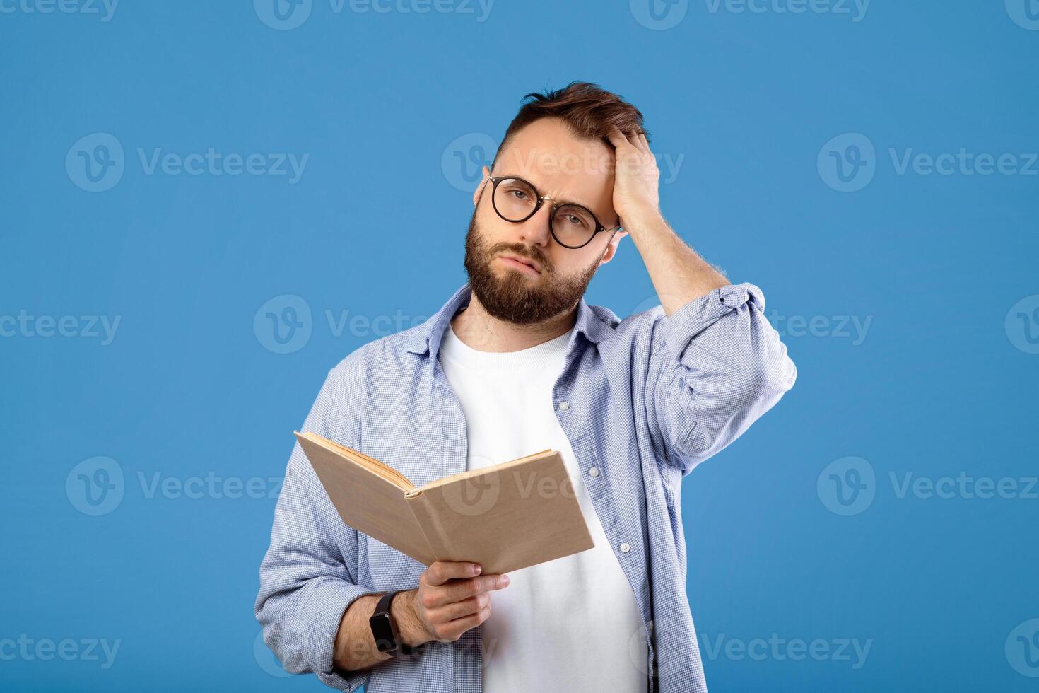 Studying is hard. Young bearded guy having problem understanding textbook on blue studio background. Millennial guy having headache while getting ready for exam or test photo