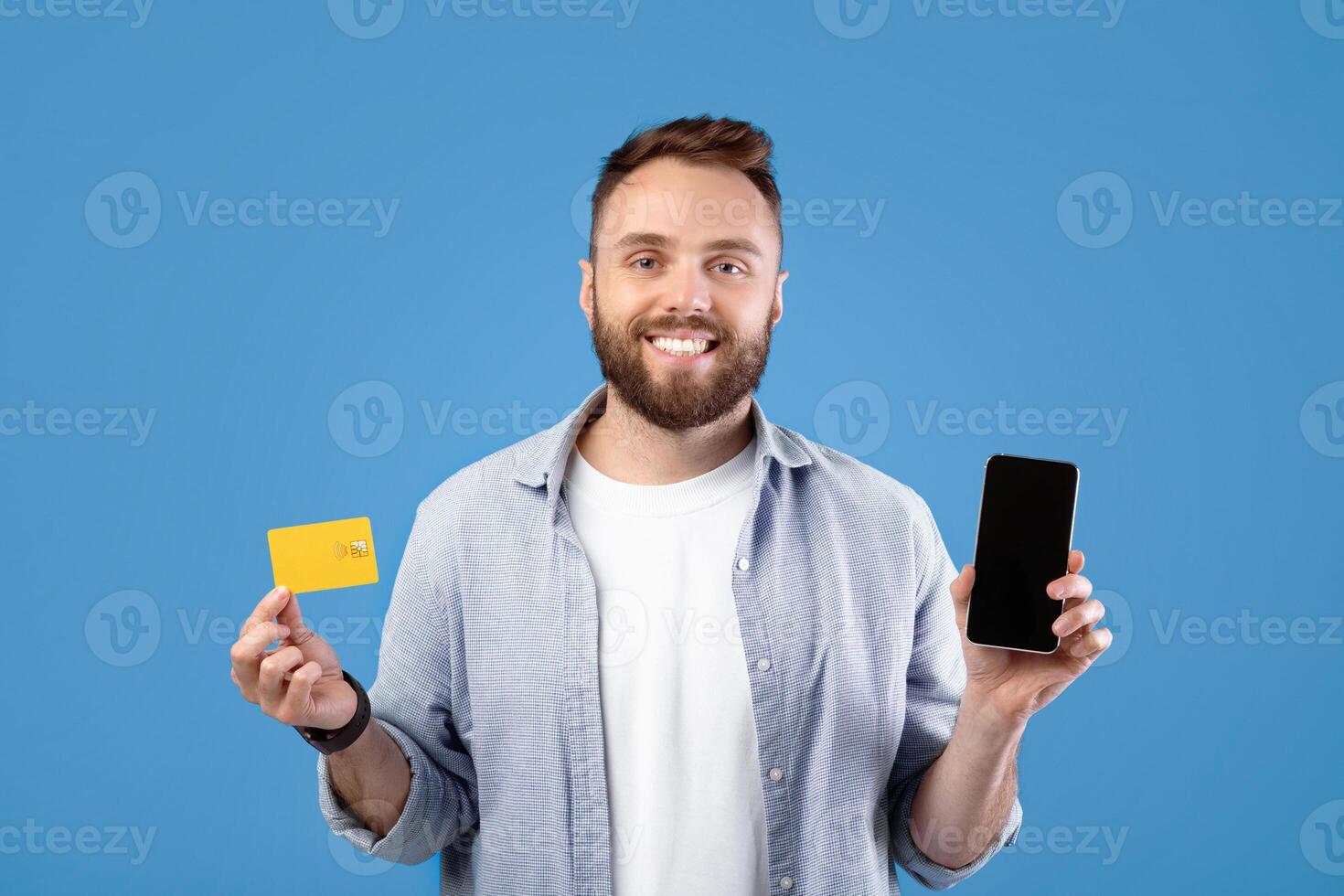 Positive millennial guy holding smartphone with empty screen and credit card on blue background, space for mobile app design. Young man promoting online shopping and contactless payment photo