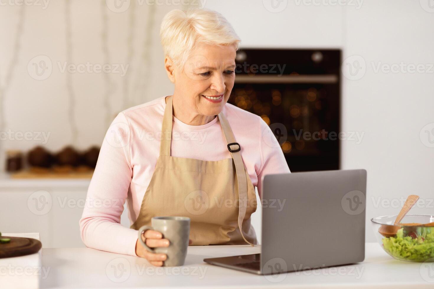 Senior Housewife Using Laptop Computer Browsing Internet For Online Recipes Cooking Dinner In Modern Kitchen At Home. Food Preparation. Retirement Hobby And Lifestyle Concept photo