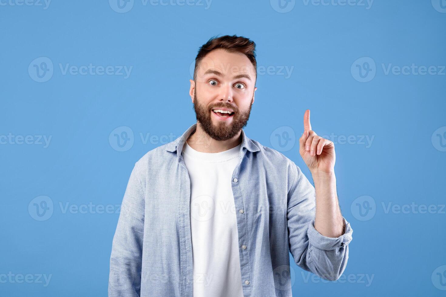 Creativity, idea, inspiration. Young handsome guy pointing finger up, gesturing eureka, experiencing AHA moment on blue studio background. Cool bearded man finding solution to difficult problem photo