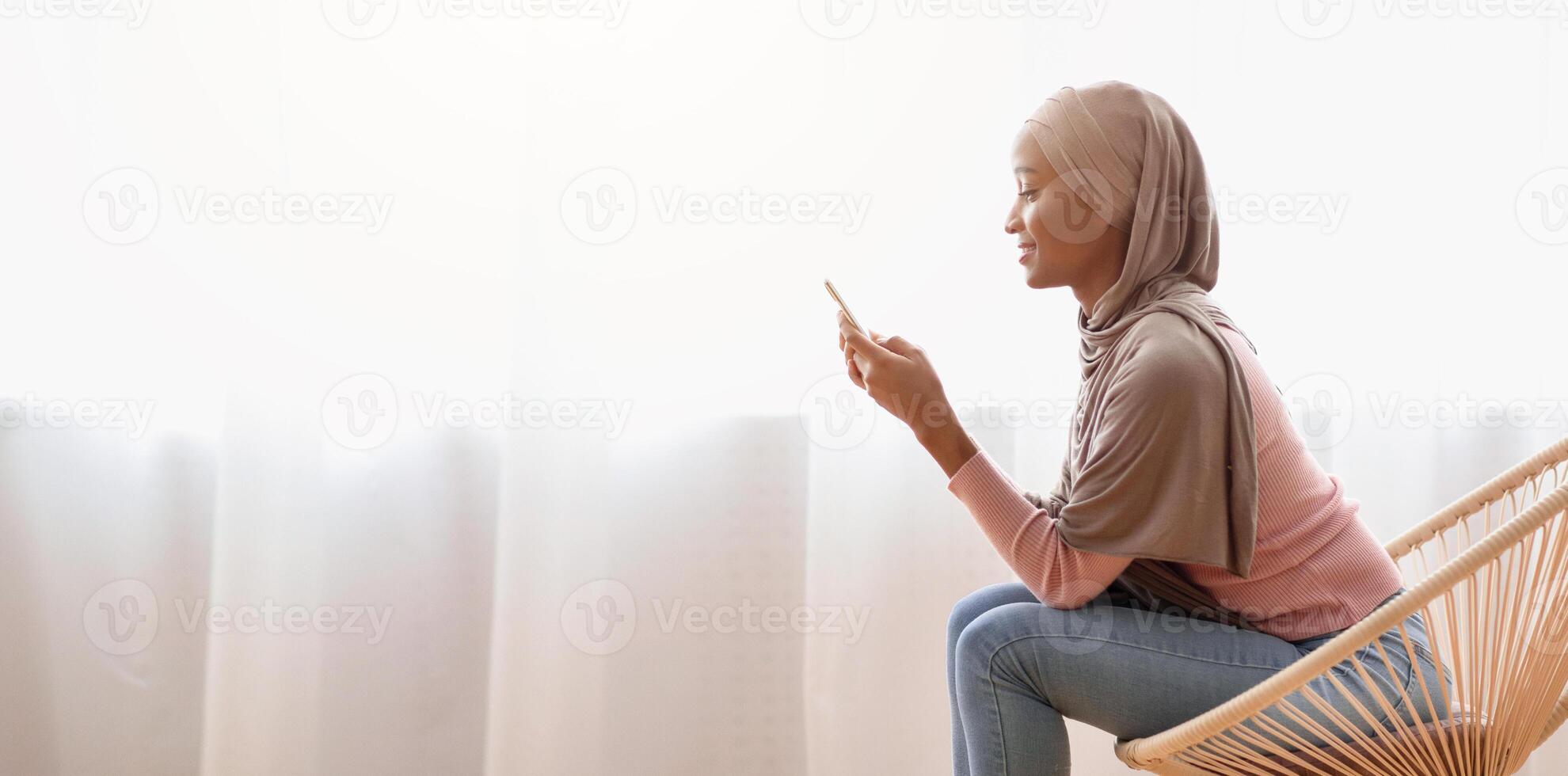 Young black Muslim woman in hijab using smartphone at home, working or studying online, copy space. African American lady in national headscarf chatting on web, taking part in conference or webinar photo